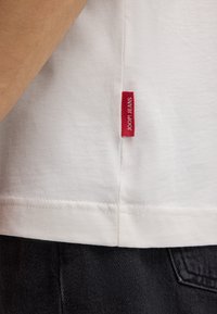 Witte katoenen t-shirt met zichtbare stiksels aan de zoom, voorzien van een rood label met "JOOP! JEANS" dat aan de zijkant is bevestigd, gecombineerd met zwarte denim.