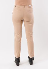 Persona che indossa pantaloni beige slim-fit e stivaletti neri, in piedi contro uno sfondo bianco semplice, vista da dietro.