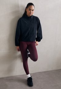 Schwarze Windjacke mit Frontreißverschluss und elastischen Bündchen, kombiniert mit burgunderfarbenen Leggings und schwarzen Sportschuhen. Einfache Kulisse.