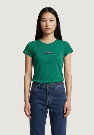 Donna con capelli neri lunghi che indossa una t-shirt verde Calvin Klein e jeans blu, in piedi davanti a uno sfondo chiaro e uniforme.