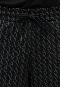 Gros plan sur un pantalon noir avec un motif répétitif gris "AX" se chevauchant sur toute la surface et un cordon noir noué en un nœud à la taille.