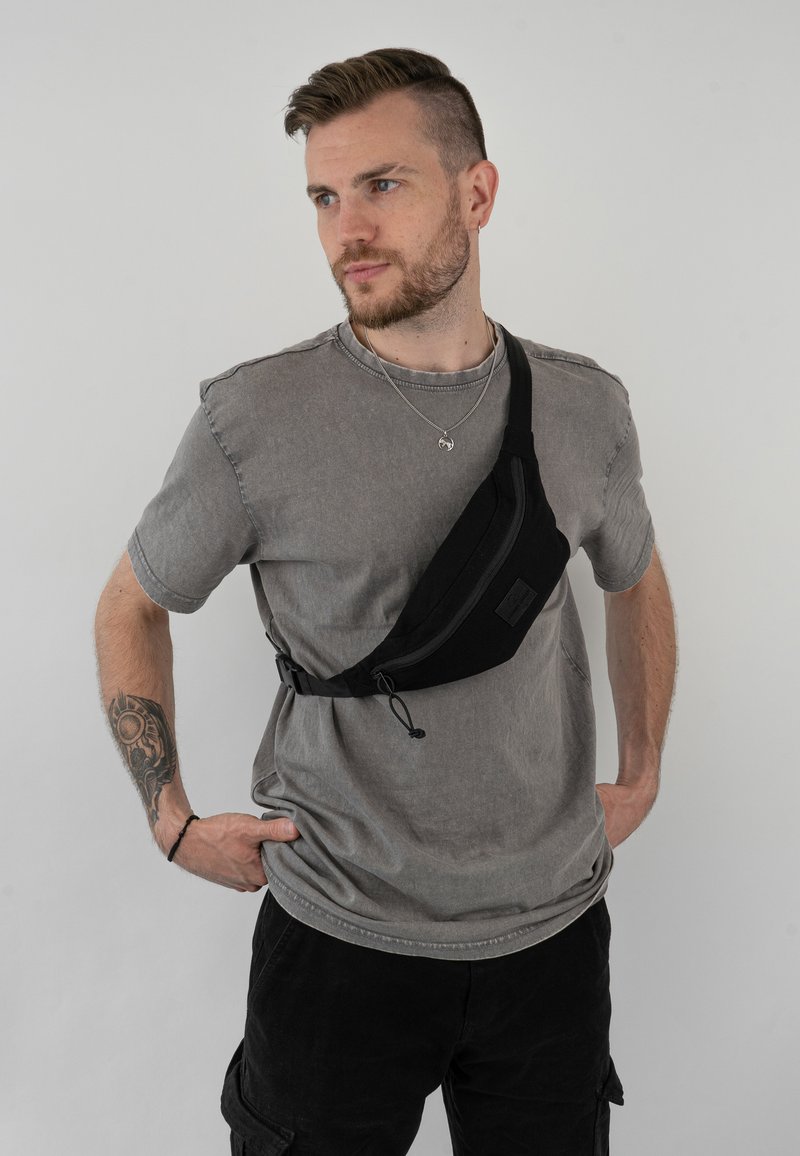 Borsa a tracolla nera in tessuto indossata sopra una maglietta grigia, con tasca con zip, tracolla regolabile e design minimalista.