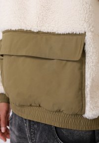 Fleecinė striukė su balta tekstūrine viršutine dalimi ir alyvuogių žalios spalvos apatine audinio dalimi. Yra didelė kišenė su atvartas ir elastinga apačia.