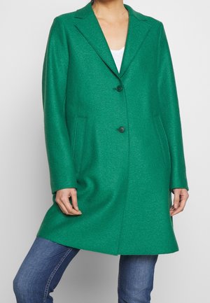 Vrouw draagt een felgroene wollen jas met twee knopen, blauwe spijkerbroek en een wit topje, staand tegen een eenvoudige lichte achtergrond.