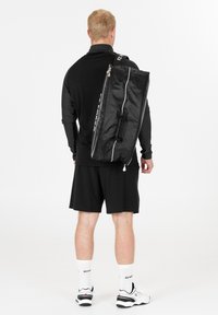 Musta urheilumatkalaukku valkoisilla yksityiskohdilla, suorakaiteen muotoinen, sileä pinta ja ylhäältä avattava vetoketju. Kuljetetaan olalla, esillä valkoista taustaa vasten.