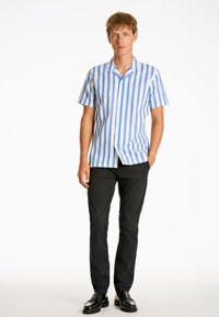 Camicia a maniche corte con bottoni, a righe verticali blu e bianche, realizzata in tessuto leggero. Abbinata a pantaloni neri e scarpe lucide.