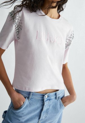 Kobieta ubrana w jasnoróżową koszulkę z krótkim rękawem, ozdobioną kamieniami na ramionach i haftowanym logo „LIU JO”, w połączeniu z jasnoniebieskimi dżinsami.