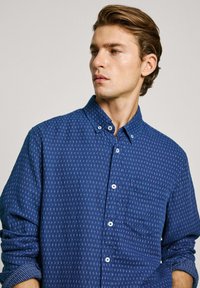 Blue button-up shirt with a small geometric pattern, long sleeves, pocket on the left side, and white buttons. Cotton fabric, casual fit.
