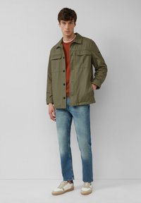 Olivgrüne Utility-Jacke mit zwei Brusttaschen, getragen über einem orangefarbenen Pullover, kombiniert mit hellblauen Jeans und weißen Sneakers.