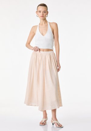 Femme portant un top court blanc à dos nu, une jupe midi beige clair et des talons ouverts, debout devant un fond blanc uni.