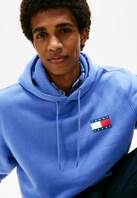 Junger Mann trägt einen blauen Tommy Jeans Hoodie mit einem karierten Hemd darunter und blickt in die Kamera vor einem schlichten weißen Hintergrund.