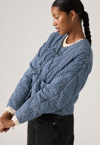 Γυναίκα με πλεξούδες, φοράει μπλε καπιτονέ καρό μπουφάν πάνω από μια κρεμ πουλόβερ, διορθώνοντας το μανίκι της, στέκεται μπροστά σε ένα σκέτο φωτεινό φόντο.