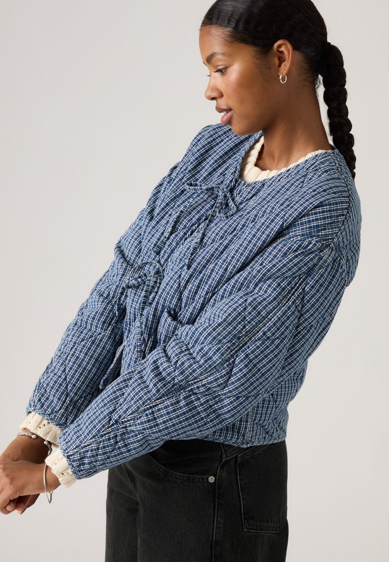 Femme aux cheveux tressés portant une veste matelassée à carreaux bleue sur un pull crème, ajustant sa manche, debout devant un fond clair uni.