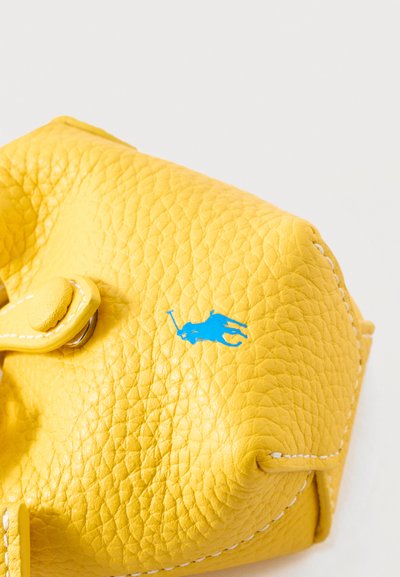 Κίτρινη γωνία δερμάτινης τσάντας με ανάγλυφη υφή, μπλε λογότυπο παίκτη πόλο και λευκό ράψιμο σε ανοιχτό φόντο.