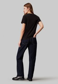 Camiseta de algodón negra con cuello redondo, combinada con jeans de mezclilla azul oscuro y botas negras; vista desde la parte trasera contra un fondo gris.