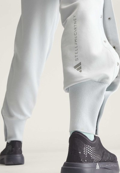 Unterschenkel mit hellen Hosen mit Stella McCartney-Logo und schwarzen Sneakern mit Mesh-Detail und Stella McCartney-Branding an der Sohle.