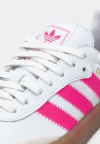 Λευκό δερμάτινο αθλητικό παπούτσι με τρεις ροζ ρίγες, υφασμάτινα κορδόνια και λογότυπο στη γλώσσα· διαθέτει υφή στην επάνω επιφάνεια και καφέ σόλα.