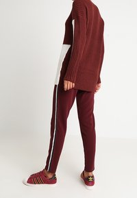 Pull en maille oversize bordeaux avec un accent blanc sur la manche, associé à un pantalon slim bordeaux avec une bande blanche latérale et des baskets sportives.