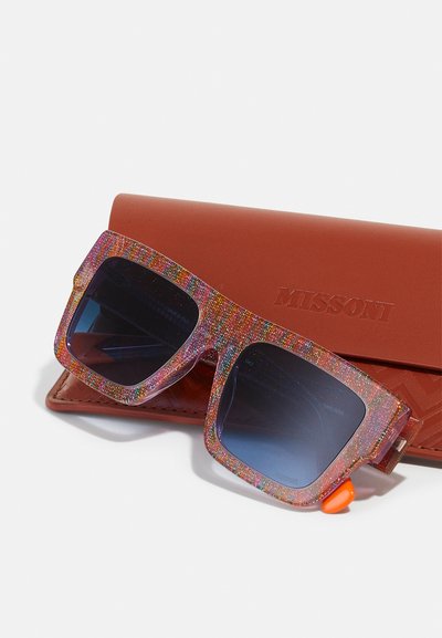 Occhiali da sole quadrati con una montatura glitterata e multicolore, lenti scure e dettagli in arancione, posizionati su una custodia in pelle marrone.