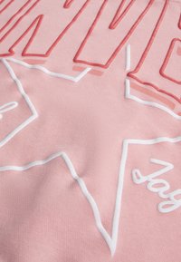 Ροζ ύφασμα με ανάγλυφα κόκκινα και λευκά γράμματα και σχέδιο αστεριού, με λεία υφή και ραμμένες λεπτομέ ρειες.