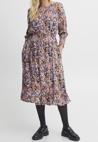 Florale midi-jurk met een paars, gele en oranje print, lange mouwen, elastische taille, zakken en een vloeiende silhouette, gecombineerd met zwarte schoenen.