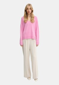 Donna che indossa un ampio maglione rosa con scollo a V, pantaloni beige larghi e scarpe beige a punta, in piedi contro uno sfondo chiaro uniforme.