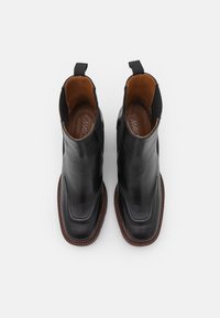 Černé kožené kotníkové boty s elegantním designem, kulatou špičkou a hnědou gumovou podrážkou. Obsahují elastické boční panely a poutka pro snadné obouvání.