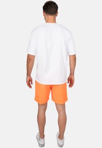 Maglietta bianca a maniche corte con scollatura rotonda abbinata a pantaloni corti arancioni vivaci. I pantaloni hanno una texture liscia e un taglio rettangolare.