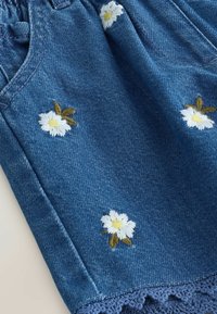 Denim rok met witte madeliefjesborduursel met gele middens en bruine bladeren. Heeft kanten trim aan de zoom in een contrasterende kleur.