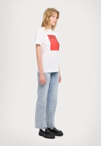 Hvit bomulls T-skjorte med et stort rødt grafisk motiv og svart tekst, sammen med lyseblå jeans og sorte kraftige plattform sko.