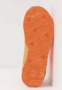 Sulan av en orange sko med ett texturerat, vågigt mönster, vilande mot en vit vägg. Varumärket "WODEN" synligt i en cirkel.