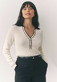 Pull en V crème avec un col noir, présentant une patte de boutonnage et des bordures noires. Porté avec un pantalon noir taille haute.
