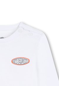 Witte langeslijf shirt met ronde halslijn, voorzien van twee drukknoopjes op de schouder en een rond Timberland-logo in oranje en zwart.