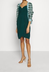 Teal Kleid mit Herz-Ausschnitt und drapiertem Design, kombiniert mit einer gemusterten Strickjacke in Hellblau und Schwarz. Tanfarbene High-Heels.