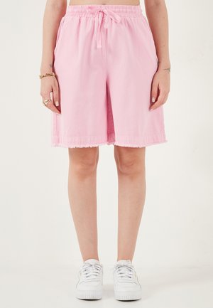 Osoba nosiaca ružové šortky sťahované šnúrkou po kolená s ošúchanými okrajmi, biele tenisky a zlaté šperky na ľavej ruke na bielom pozadí.