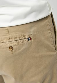 Gros plan sur la poche arrière d'un pantalon beige avec bouton et petit détail en tissu rouge, blanc et bleu, associé à une chemise blanche.