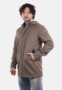 Giacca marrone con cappuccio, con texture a coste, tasche con zip e un logo circolare sulla manica. Presenta una vestibilità rilassata e un cappuccio regolabile.