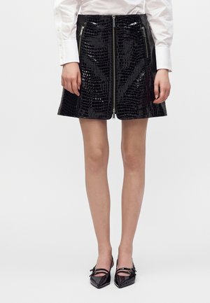 Femme portant une mini-jupe noire brillante à texture crocodile avec fermeture éclair devant et poches zippées, chemise blanche à manches longues et chaussures noires pointues.
