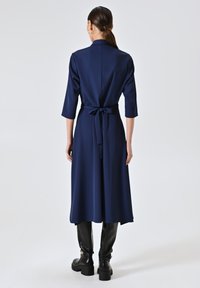Marineblauwe jurk tot de knie met een aansluitend lijfje, wijde mouwen en een ceintuur op de taille. Glad materiaal met een split aan de achterkant en minimale details.