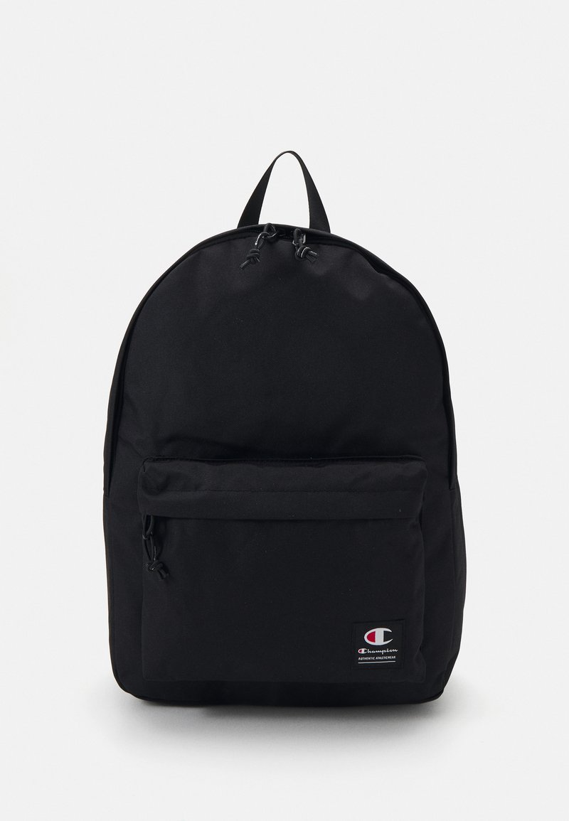 Sac à dos noir en tissu durable, présentant une forme arrondie, une poignée supérieure, une poche zippée à l'avant et un patch logo dans le coin inférieur.