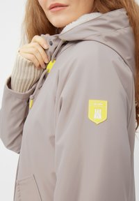 Lichtbeige regenjas met een fleece-voering in de kraag, gele accenten en een schildlogo op de mouw. Bevat een voorzak en een capuchon met koord.