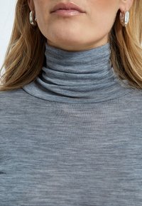 Šedý rolák s jemnou, textúrovanou tkaninou, ktorý má priliehavý strih a vrstvený golier; doplnený lesklými kruhovými náušnicami.