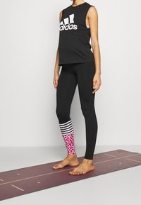 Osoba stojící bosky na jóga podložce, oblečená v černém bezrukávovém tričku Adidas a černých legínách se pruhovaným a růžovým leopardím vzorem u kotníků.