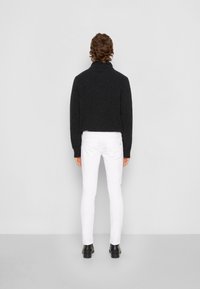 Suéter de punto gris oscuro con cuello alto, combinado con pantalones ajustados blancos y botines negros, visto desde la parte de atrás.