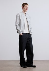 Ανοιχτό γκρι μπουφάν bomber με ριμπ μανσέτες, συνδυασμένο με σκούρα μπλε τζιν παντελόνια με φαρδύ πόδι. Το μοντέλο στέκεται με τα χέρια στις τσέπες μπροστά σε ουδέτερο φόντο.