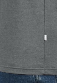 T-shirt in tessuto grigio con una texture morbida, caratterizzato da un orlo cucito e un piccolo etichetta bianca sulla cucitura laterale. Indossato con jeans in denim blu.