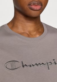 Gray cotton t-shirt featuring an embroidered "Champion" logo in darker gray. Round neckline with a smooth texture, close-up view.