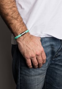 Bracciale intrecciato in turchese con una chiusura metallica, indossato al polso, che mostra un design texturizzato su una mano e uno sfondo in denim.