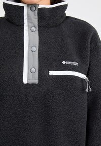 Schwarzer Fleece-Pullover mit hohem Kragen, grauem Druckknopfverschluss und weißen Akzenten. Verfügt über eine zip-fähige Brusttasche und das Columbia-Logo. Weiche Textur.
