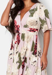 Femme portant une robe portefeuille rose à motifs floraux, avec de grandes fleurs blanches, rouges et vertes, des manches courtes et une taille cintrée.